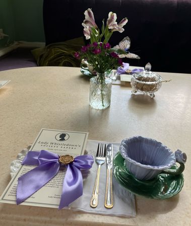 Afternoon tea table in a cozy tea room: lavender-ribboned menu, gold-trimmed fork and knife, scalloped lavender teacup on a green saucer, crystal sugar bowl, and small vase of pink and white flowers with a decorative butterfly.