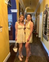 Cheerful scene of two women in light yellow summer dresses smiling and posing in a decorative indoor hallway with yellow walls, ornate silver mirror, arched blue doorway, and hardwood floors.