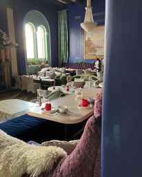 Cozy vintage tea room interior with deep blue walls and arched windows, crystal chandelier, tufted purple banquette, plush mismatched seating and tables set for afternoon tea with teacups and red napkin roses.