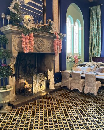 Ornate Victorian-style tea room interior with carved stone fireplace draped in pink floral garlands, lit fire, arched mint-green windows, velvet dining chairs and patterned carpet.