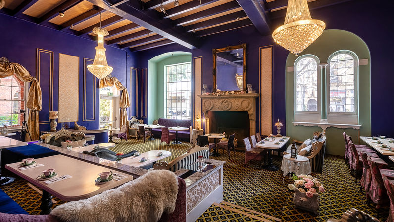 Elegant purple Victorian-style tea room interior with crystal chandeliers, ornate fireplace, arched windows, vintage seating and tables set with teacups and floral accents.
