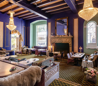 Elegant purple Victorian-style tea room interior with crystal chandeliers, ornate fireplace, arched windows, vintage seating and tables set with teacups and floral accents.