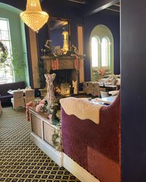Cozy vintage tea room interior with glowing crystal chandelier, ornate fireplace draped in floral garlands, plush velvet seating and small tea tables by arched windows on a patterned carpet