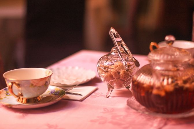 Vintage floral porcelain teacup and saucer, glass sugar jar with tongs, and amber glass teapot on a pink tablecloth — cozy afternoon tea table setting.