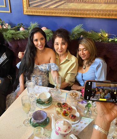 Three smiling women in a cozy blue-walled tea room pose at a table set with a floral teapot, teacups, champagne flutes and assorted pastries while a phone captures their photo; garland and gold-framed painting behind them.
