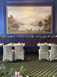 Cozy boutique tea room interior with a royal-blue wall and oversized gold-framed lakeside landscape painting, tufted plum banquette, floral garland, patterned carpet, and small tables with slipcovered chairs set for tea.