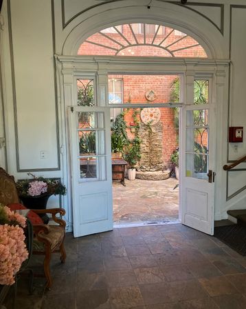 View through white arched double doors into a sunlit brick courtyard with a stacked-stone fountain, potted plants, and a vintage chair with pink flowers.