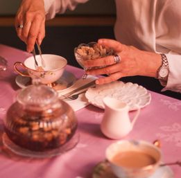 Close-up of hands using tongs to add brown sugar cubes to a floral teacup on a pink tablecloth, with a glass sugar bowl, white creamer, saucer and a filled teacup nearby.