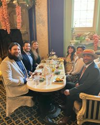 Six people in semi-formal attire enjoying afternoon tea at a small round table in an ornate Victorian-style tearoom, with tiered pastries, teapots, floral garlands and a sunlit window.