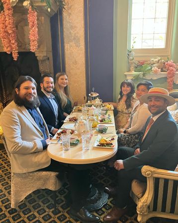 Six people in semi-formal attire enjoying afternoon tea at a small round table in an ornate Victorian-style tearoom, with tiered pastries, teapots, floral garlands and a sunlit window.