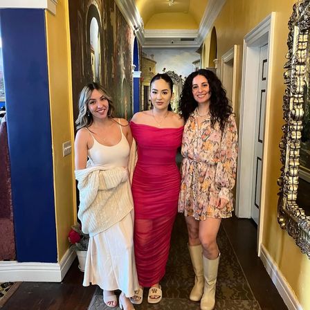 Three women in summer dresses smiling and posing in a colorful restaurant hallway with yellow walls, ornate mirror, painted mural and wood floors