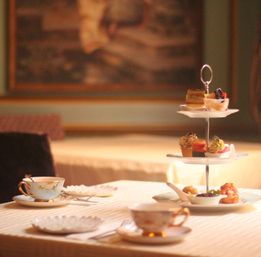 Elegant afternoon tea spread in a cozy tearoom: fine china teacups and a three-tier stand with pastries, scones, and savory bites.
