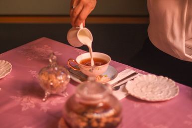 Elegant vintage tea service — milk poured into a floral porcelain teacup on a pink damask tablecloth, with a silver sugar bowl and scalloped plate.