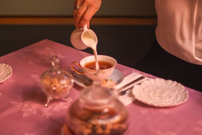 Elegant vintage tea service — milk poured into a floral porcelain teacup on a pink damask tablecloth, with a silver sugar bowl and scalloped plate.