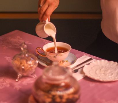Elegant vintage tea service — milk poured into a floral porcelain teacup on a pink damask tablecloth, with a silver sugar bowl and scalloped plate.