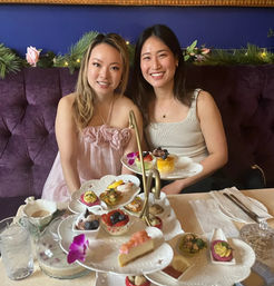 Two smiling friends seated on a purple velvet banquette enjoying afternoon tea with a three-tiered stand of colorful pastries, finger sandwiches, fresh fruit and floral accents.