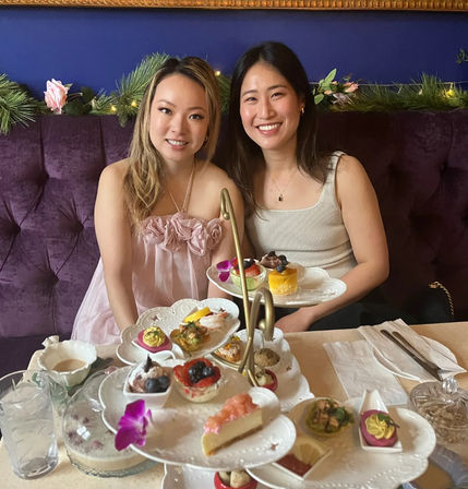 Two smiling friends seated on a purple velvet banquette enjoying afternoon tea with a three-tiered stand of colorful pastries, finger sandwiches, fresh fruit and floral accents.