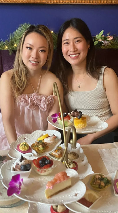 Two smiling friends seated on a purple velvet banquette enjoying afternoon tea with a three-tiered stand of colorful pastries, finger sandwiches, fresh fruit and floral accents.