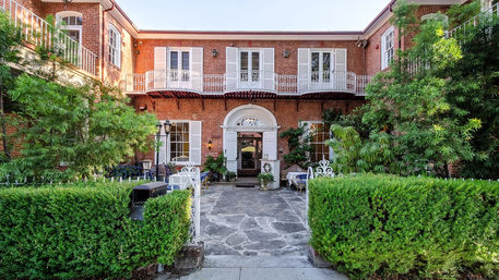 Sunlit red-brick two-story building with white shutters and decorative wrought-iron balcony, stone courtyard entrance flanked by manicured boxwood hedges, outdoor seating and lush greenery.