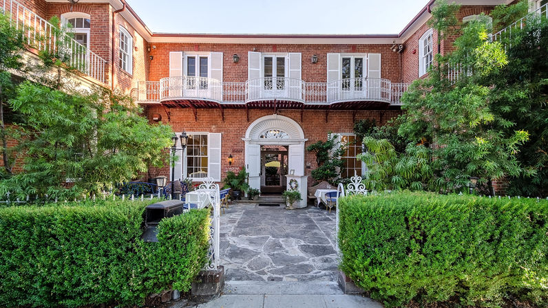 Sunlit red-brick two-story building with white shutters and decorative wrought-iron balcony, stone courtyard entrance flanked by manicured boxwood hedges, outdoor seating and lush greenery.