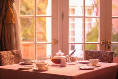 Cozy sunlit afternoon tea scene with a glass teapot, porcelain teacups and saucers on a pink tablecloth at a small dining table by French doors overlooking a garden.