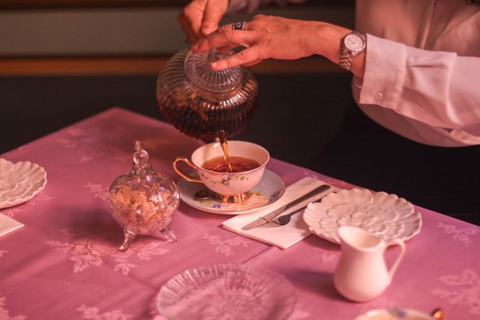 Cozy afternoon tea scene with hands pouring amber tea from a ribbed glass teapot into a floral porcelain teacup on a pink patterned tablecloth, beside a crystal sugar jar, white creamer, plates and cutlery.