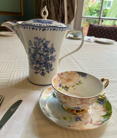 Blue-and-white floral porcelain teapot and gold-rimmed floral teacup with matching saucer on a white tablecloth at an indoor tea table by a window
