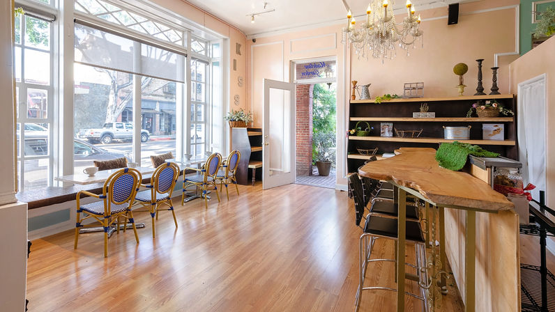 Sunlit, charming café interior with floor-to-ceiling street-facing windows, blue bistro chairs at a window bar, warm wooden floors, a live-edge wooden counter with metal stools, decorative shelving and a chandelier, open door to the sidewalk.