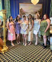 Bridal shower group: six women in pastel floral dresses with the bride in a veil and sash, posed in an ornate blue parlor with chandelier, harp, and patterned carpet.