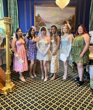 Bridal shower group: six women in pastel floral dresses with the bride in a veil and sash, posed in an ornate blue parlor with chandelier, harp, and patterned carpet.