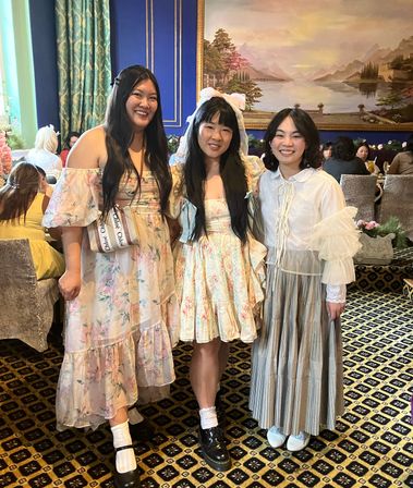 Three smiling women in pastel floral and lace dresses pose together inside an elegant tea-room with patterned carpet, velvet chairs and a large landscape painting.