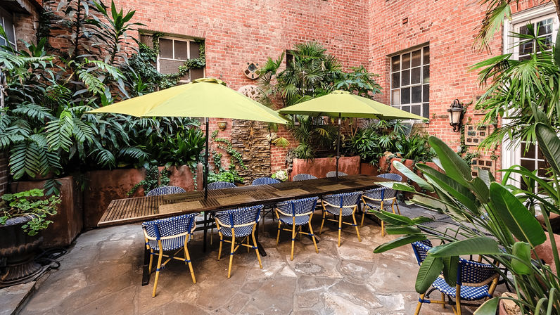 Urban brick-walled courtyard with a long wooden dining table, lime-green umbrellas and blue woven bistro chairs on a stone patio, surrounded by lush potted tropical plants