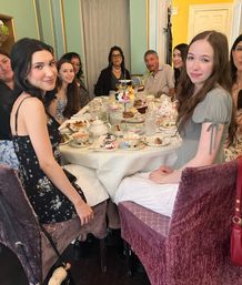 Smiling group gathered around a lace-covered table in a cozy vintage tearoom enjoying afternoon tea with tiered pastry stand, teapots and desserts