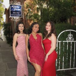 Three friends in red and pink evening gowns smiling and posing by a white wrought-iron garden gate outside a brick building on a tree-lined sidewalk