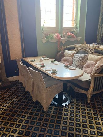 Cozy vintage tea room seating by a sunlit window with pastel teacups on a long table, ornate carved sofa, and floral window decor