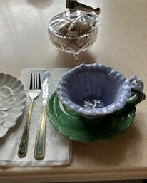 Polished table setting on a cream countertop with a fluted lavender teacup featuring a rose handle on a green saucer, gold-trimmed fork and knife on a folded white napkin, and a cut-crystal sugar bowl with tongs.