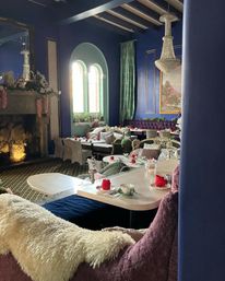 Cozy, elegant tearoom interior with deep blue walls and arched windows, crystal chandelier and fireplace, tufted velvet banquettes, plush throws, and tables set for afternoon tea with teacups and red napkins.