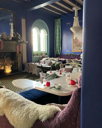 Cozy, elegant tearoom interior with deep blue walls and arched windows, crystal chandelier and fireplace, tufted velvet banquettes, plush throws, and tables set for afternoon tea with teacups and red napkins.