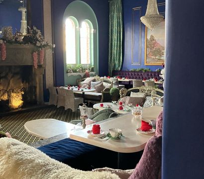 Cozy, elegant tearoom interior with deep blue walls and arched windows, crystal chandelier and fireplace, tufted velvet banquettes, plush throws, and tables set for afternoon tea with teacups and red napkins.