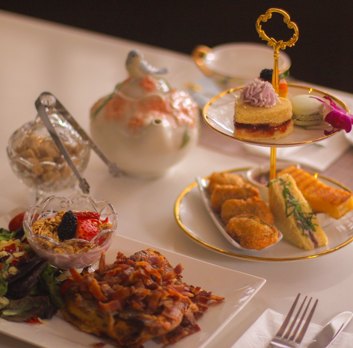 Afternoon tea spread with a gold-rimmed two-tier stand holding finger sandwiches, pastries and a macaron, a floral porcelain teapot, crystal bowls of granola and berries, and a bacon-topped savory plate on a white table.