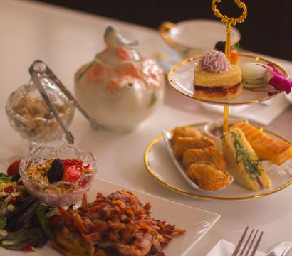 Afternoon tea spread with a gold-rimmed two-tier stand holding finger sandwiches, pastries and a macaron, a floral porcelain teapot, crystal bowls of granola and berries, and a bacon-topped savory plate on a white table.