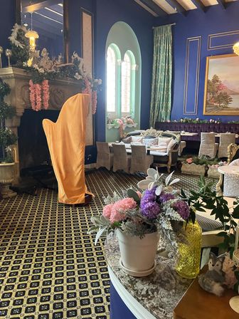 Elegant European-style lounge with deep blue walls, arched windows, patterned carpet and plush velvet seating; carved fireplace with floral garlands, a golden-draped object, and potted pink and purple flowers in the foreground.