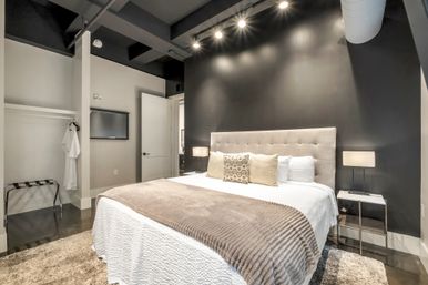 Sleek modern loft bedroom with a tufted king bed against a black accent wall, neutral pillows and textured throw, bedside lamps, track lighting, open closet and polished concrete floor