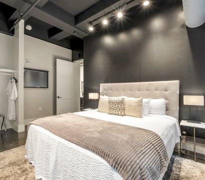 Sleek modern loft bedroom with a tufted king bed against a black accent wall, neutral pillows and textured throw, bedside lamps, track lighting, open closet and polished concrete floor