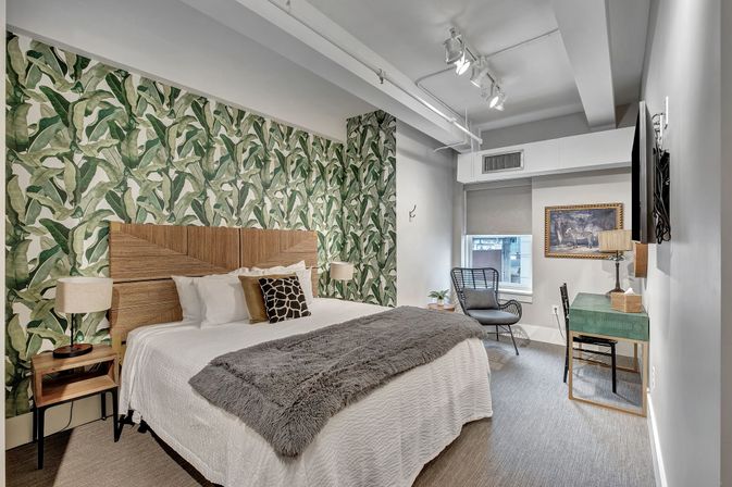 Stylish modern bedroom with tropical green leaf wallpaper, king bed with wooden headboard and gray faux-fur throw, matching bedside lamps, wicker lounge chair by a city window, and a small desk with wall-mounted TV.