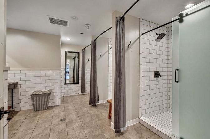 Modern communal shower room with white subway tile, gray curtains, frosted glass door, bench and full-length mirror on a tiled floor