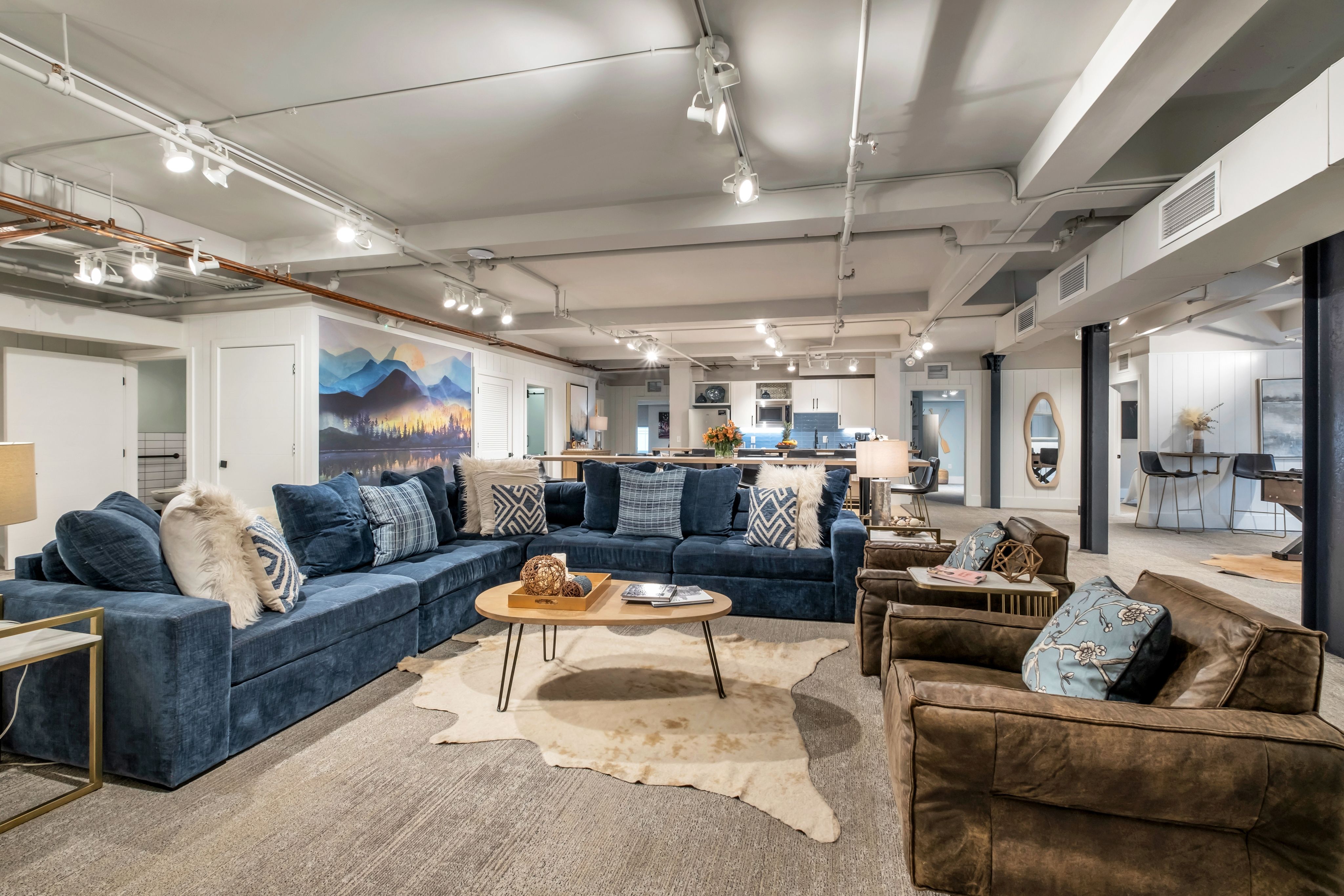 Cozy open-concept finished basement lounge with a large navy sectional, brown leather armchairs, wooden coffee table on a cowhide rug, mountain mural on the wall and a modern kitchen in the background under track lighting.