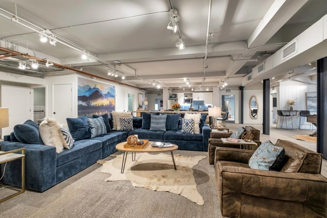 Cozy open-concept finished basement lounge with a large navy sectional, brown leather armchairs, wooden coffee table on a cowhide rug, mountain mural on the wall and a modern kitchen in the background under track lighting.