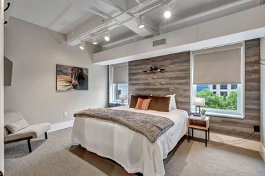 Cozy modern urban loft bedroom with wood accent wall, queen bed in white bedding and faux fur throw, brown and striped pillows, bedside table with lamp and plant, two windows with roller shades showing city greenery, track lighting, horse artwork and upholstered lounge chair.