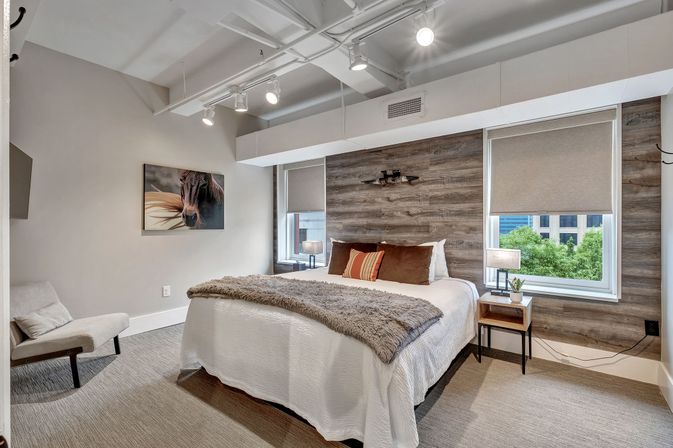Cozy modern urban loft bedroom with wood accent wall, queen bed in white bedding and faux fur throw, brown and striped pillows, bedside table with lamp and plant, two windows with roller shades showing city greenery, track lighting, horse artwork and upholstered lounge chair.